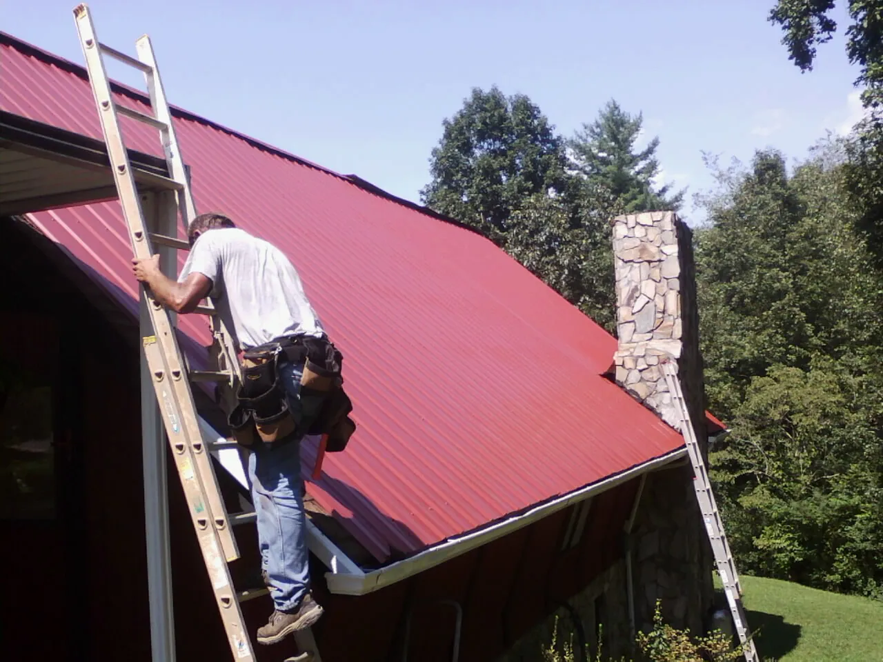 Roofer on a ladder beside a red metal roof and stone chimney — Soffit & Fascia Repair by Roofing Brothers in Alvin